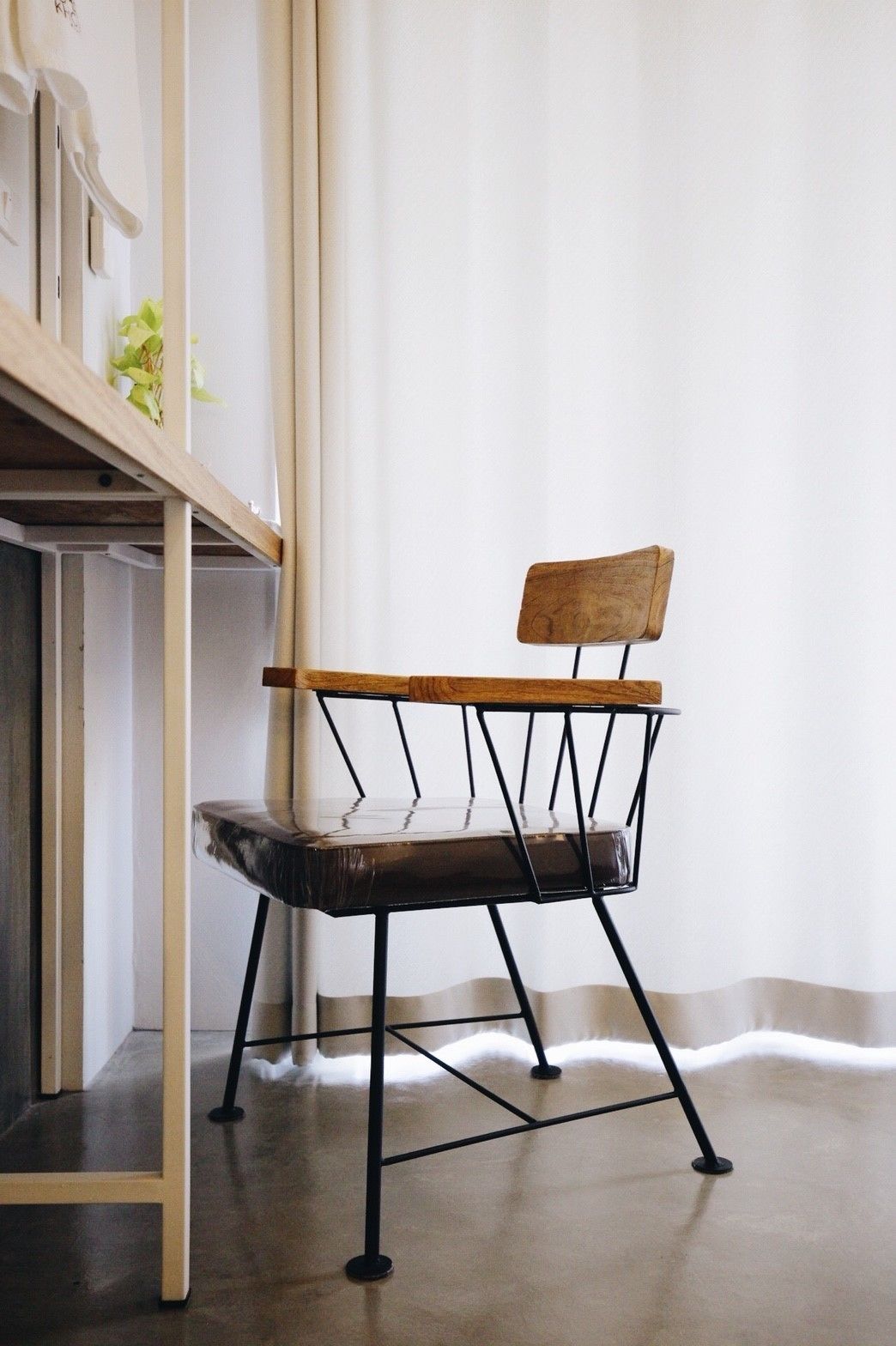 Una silla con respaldo de madera y estructura metálica, ubicada en una habitación con pared blanca y una estantería de madera con objetos encima.