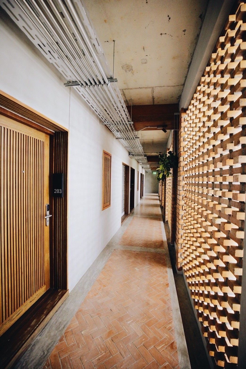 Un pasillo con una puerta de madera que lleva la etiqueta '203', una pared de ladrillo con un diseño geométrico y un piso de baldosas.