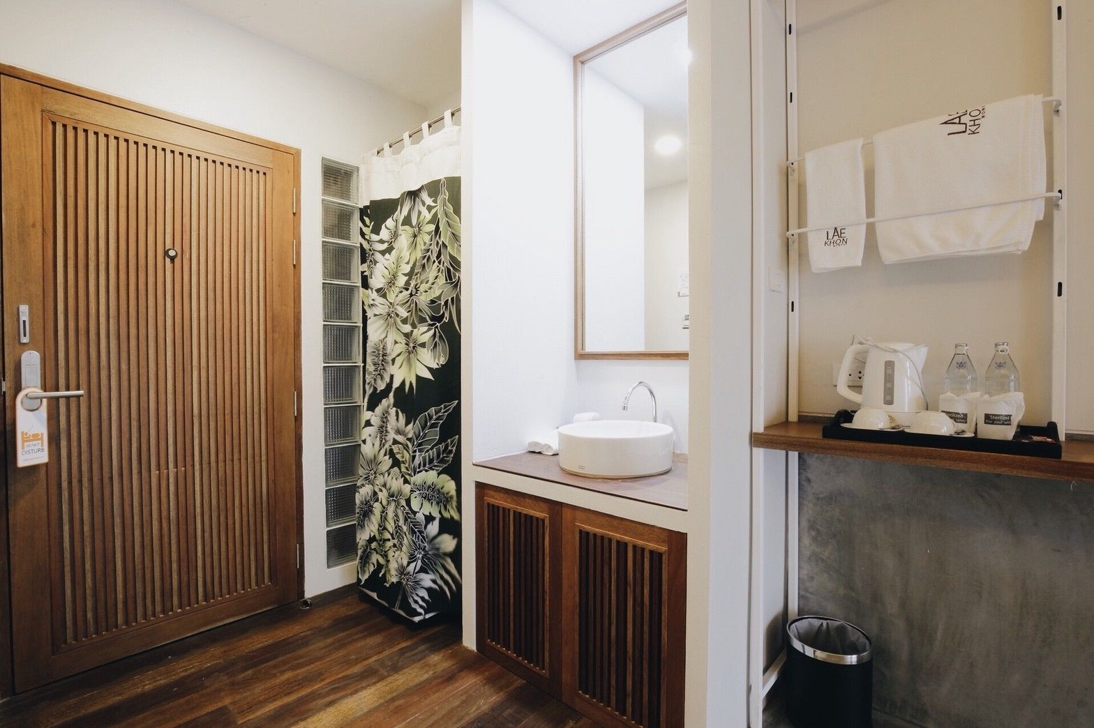 A modern bathroom with a wooden door, a floral shower curtain, a sink with a mirror, and a shelf with towels and toiletries.