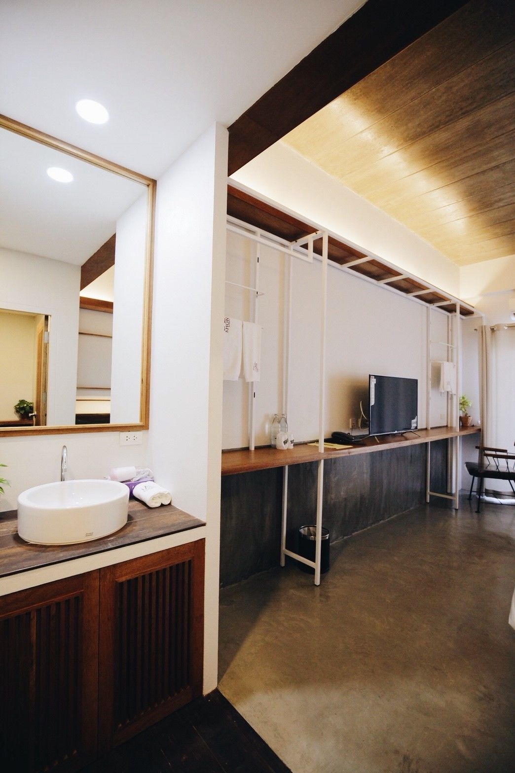 Un baño moderno con un espejo grande, un lavamanos y un mueble de tocador. Hay un televisor en una estantería y un cubo de basura.