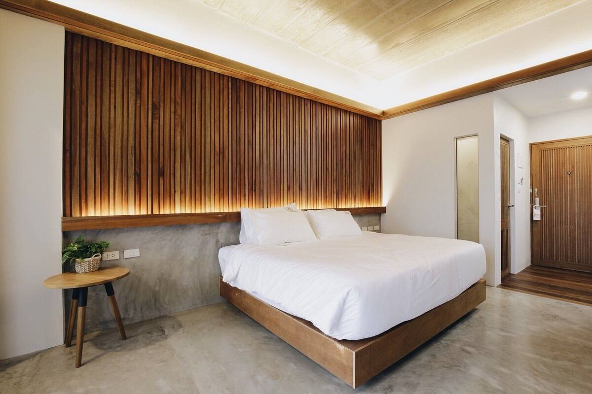 Un dormitorio moderno con cabecera de madera, una cama con ropa blanca, una mesita redonda con una planta y una puerta de madera.