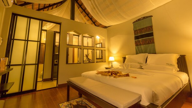 A hotel room with a large bed, a bench, a bedside table with a lamp, and a wall with a patterned tapestry. - Pou Villa (Luang Namtha)
