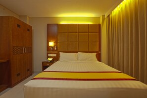 A hotel room with a large bed, a wooden wardrobe, a nightstand with a lamp, and a headboard with a light panel.