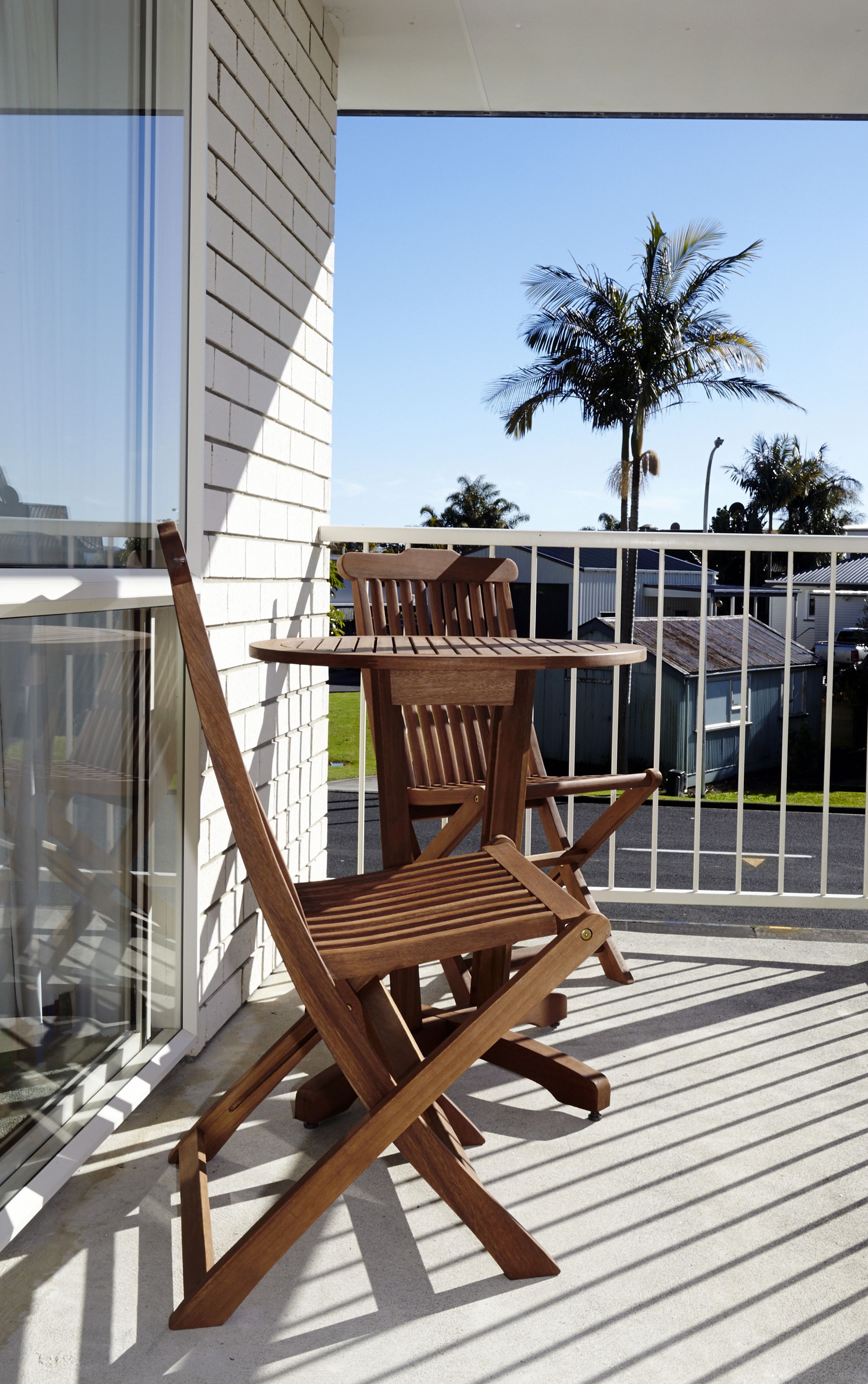 A wooden outdoor table and chairs on a balcony with a glass railing, overlooking a residential area with palm trees and houses.