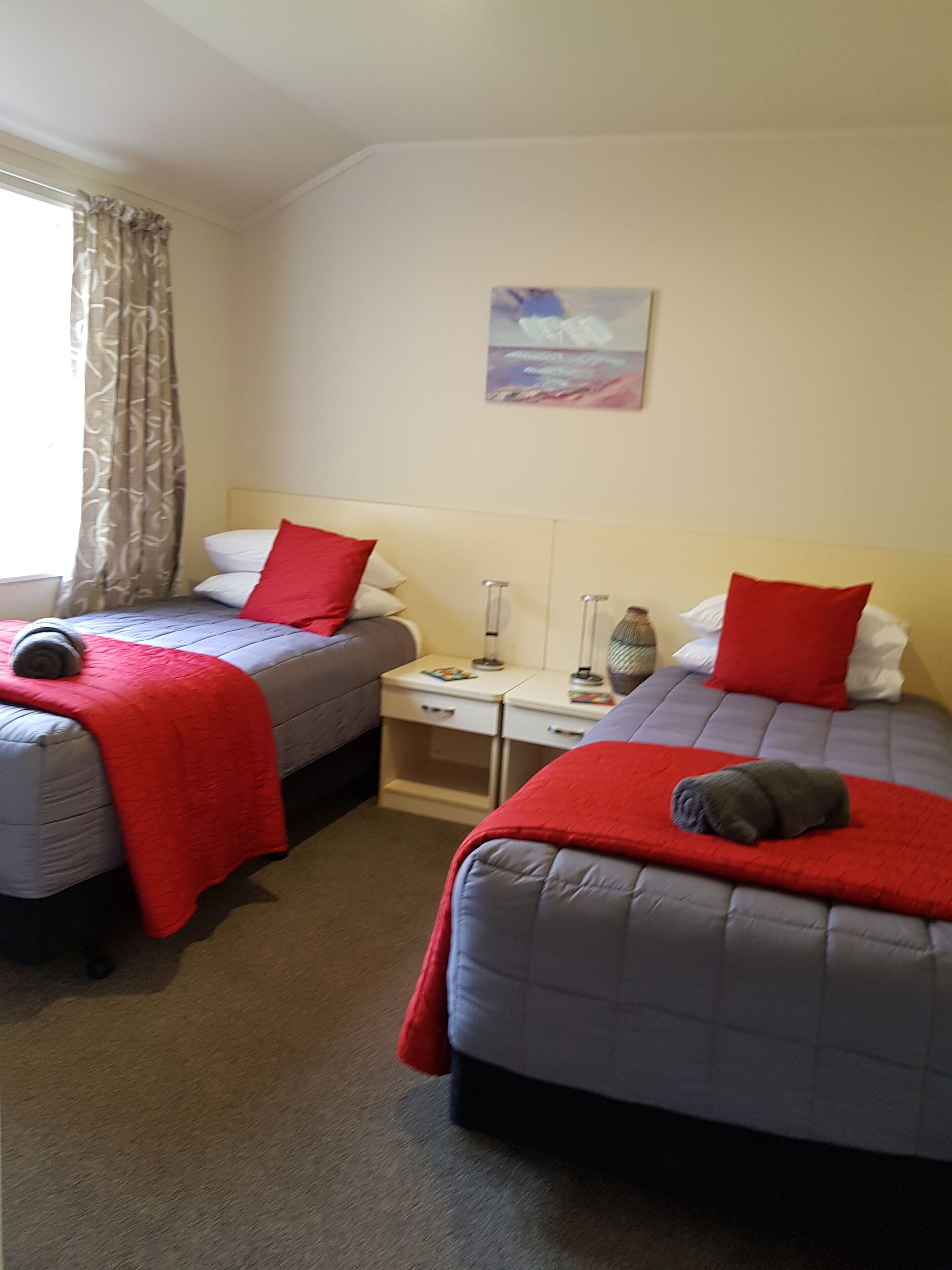A hotel room with two beds, each with a red blanket and pillow, a bedside table with a lamp, and a window with curtains.