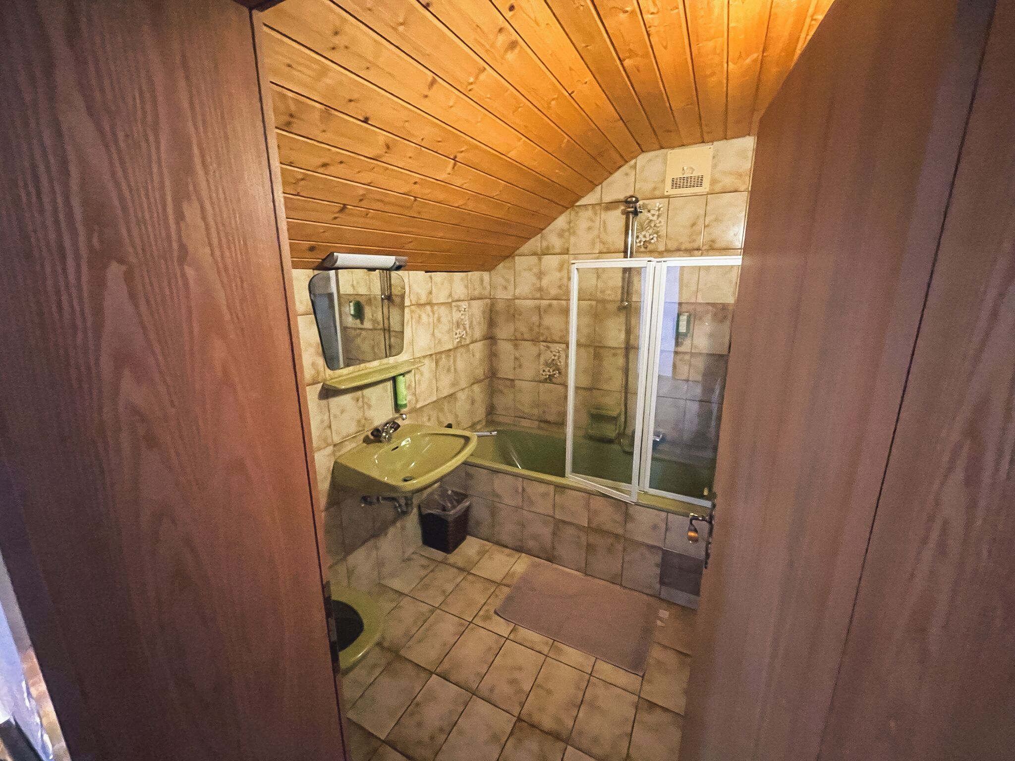 Un baño pequeño y rústico con techo de madera, un lavabo verde, un espejo, cabina de ducha y suelo de baldosas.