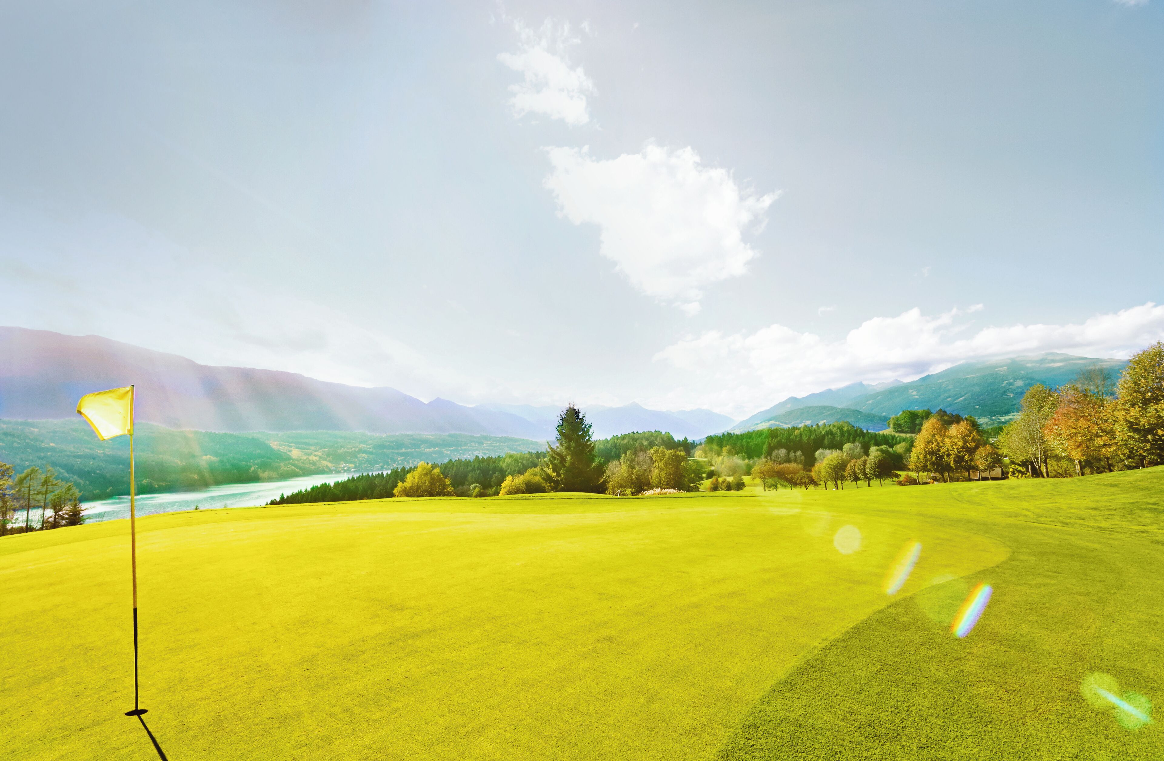 A golf course with a flagstick in the foreground, surrounded by a lush green landscape and distant mountains.