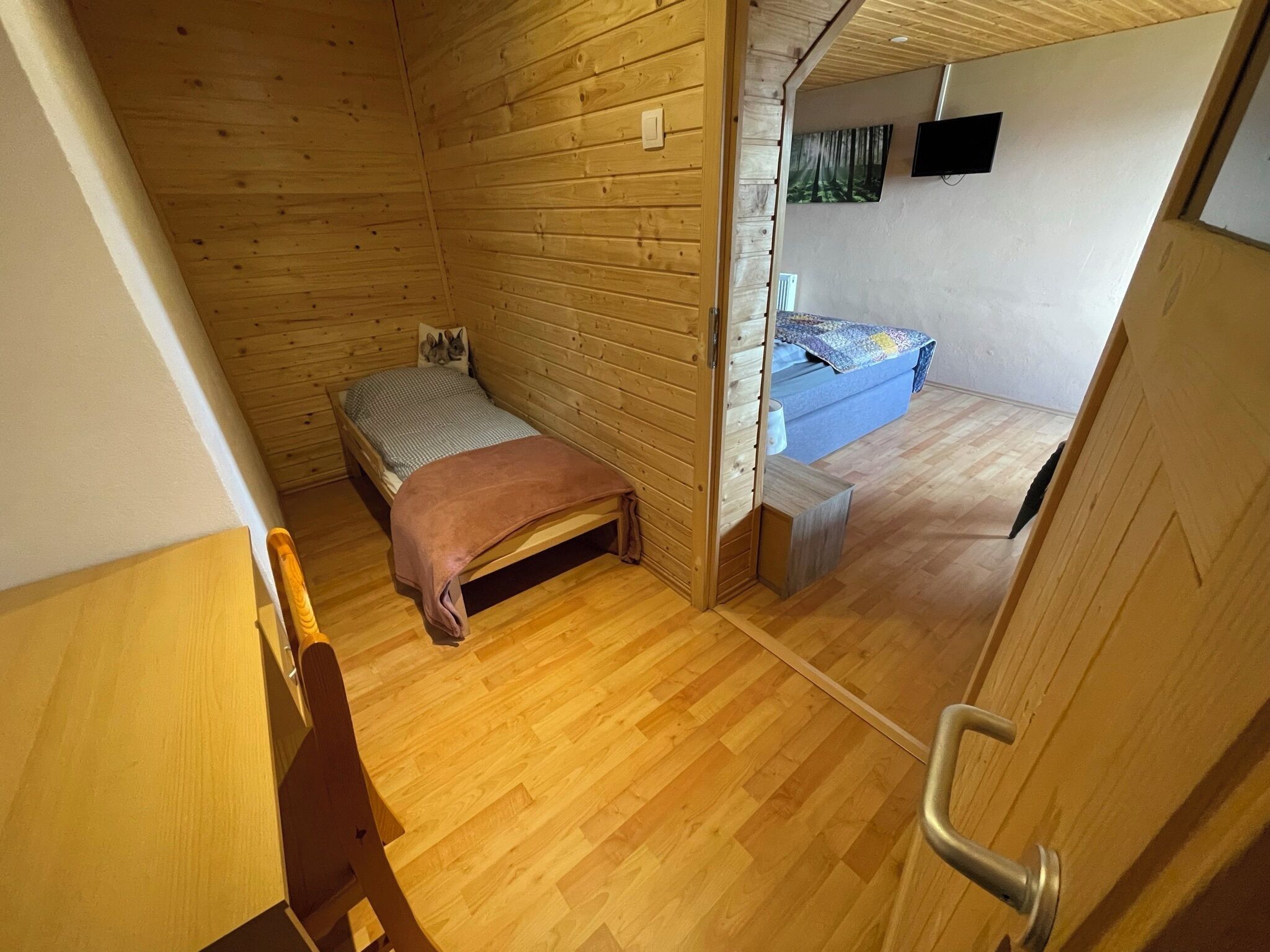 Et lille værelse med trægulv, en enkel seng, et lille bord og en stol.