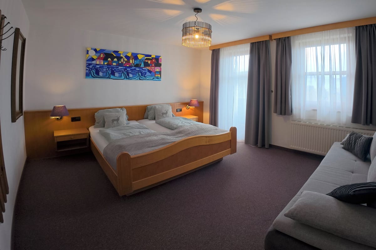Une chambre d’hôtel avec un lit en bois, un canapé, un tableau au mur et une fenêtre avec des rideaux.