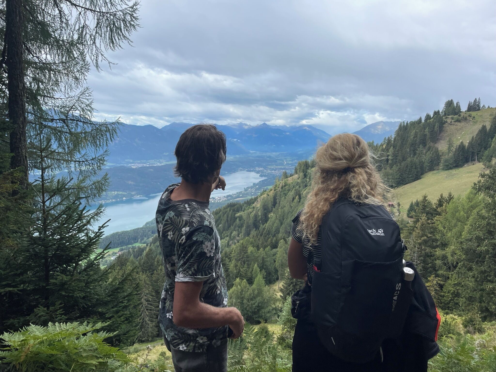 Two people standing on a mountain overlooking a valley with a lake and mountains in the distance.