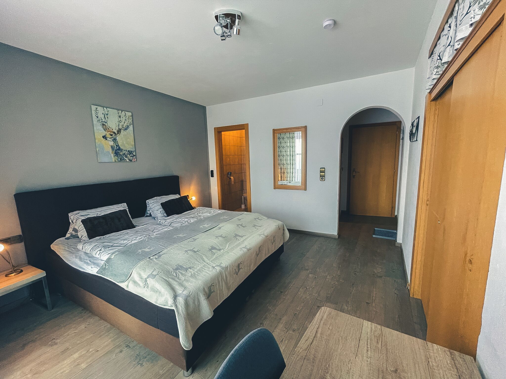 A bedroom with a large bed, a nightstand, a desk, and a chair. There are two doors and a window.