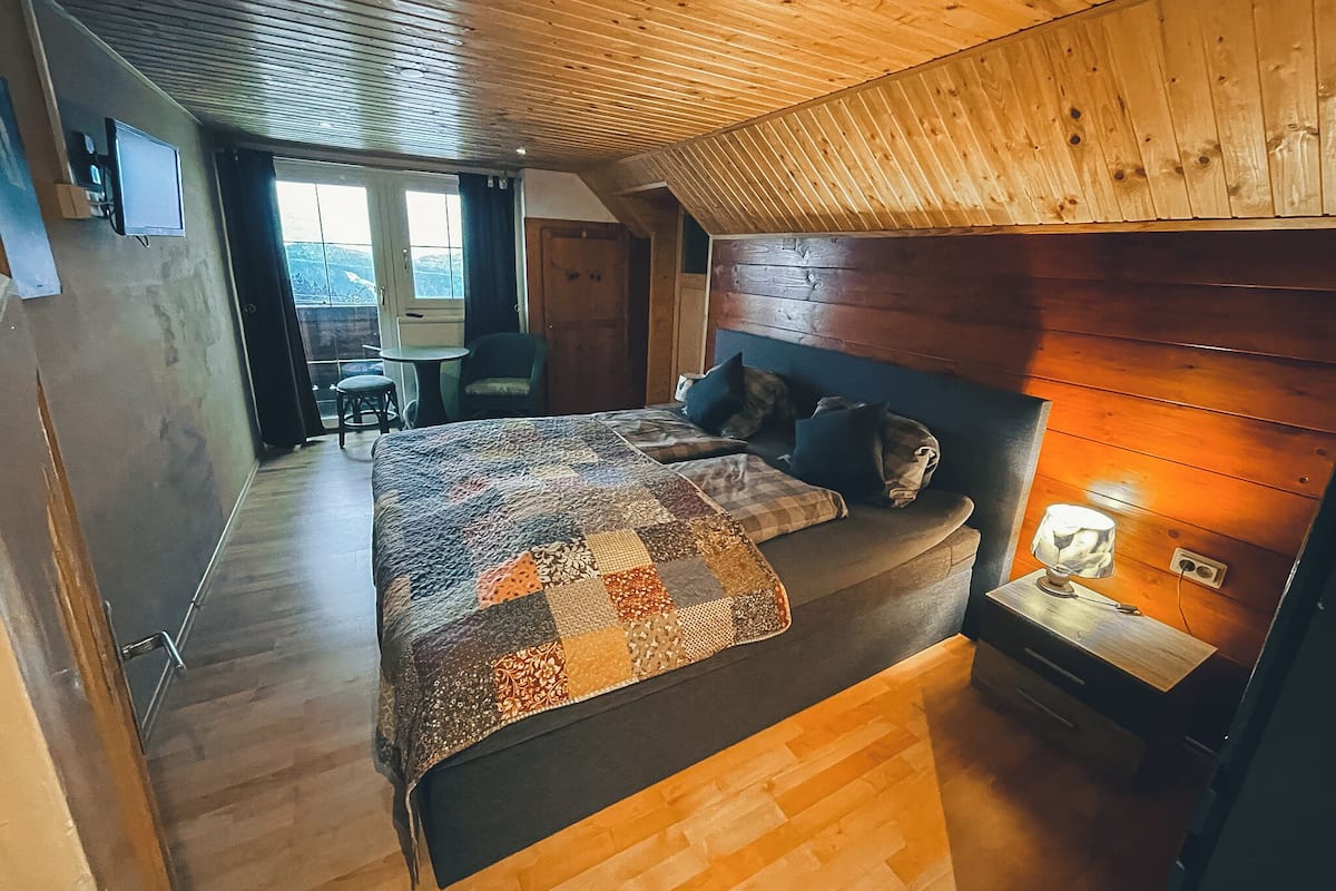 Une chambre confortable avec un plafond en bois, un grand lit recouvert d’une courtepointe, une table de chevet avec une lampe et une vue sur l’extérieur par une fenêtre.