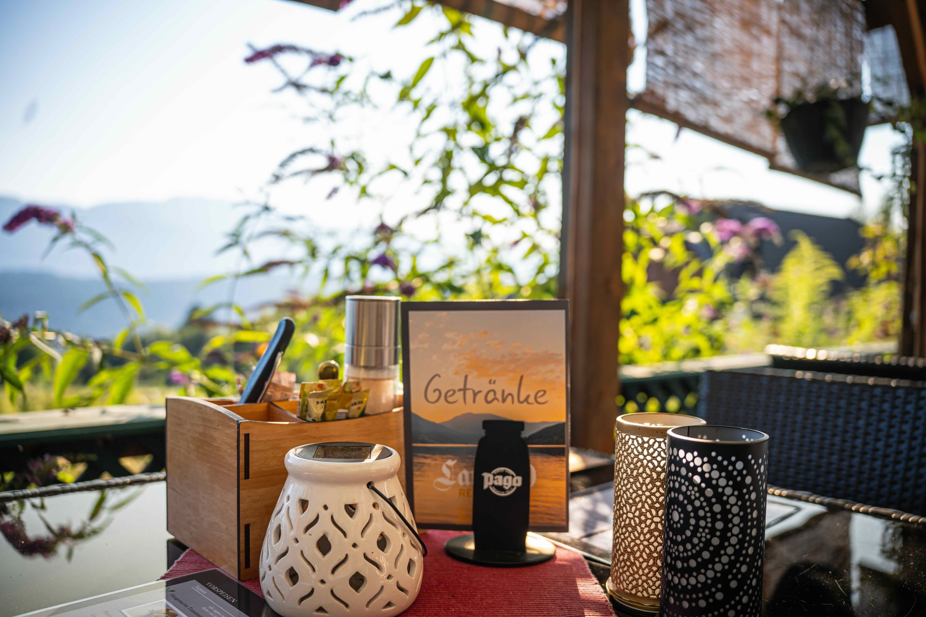 A table with a wooden box, a white vase, a black bottle, and two patterned candles, set against a backdrop of greenery and a scenic view.