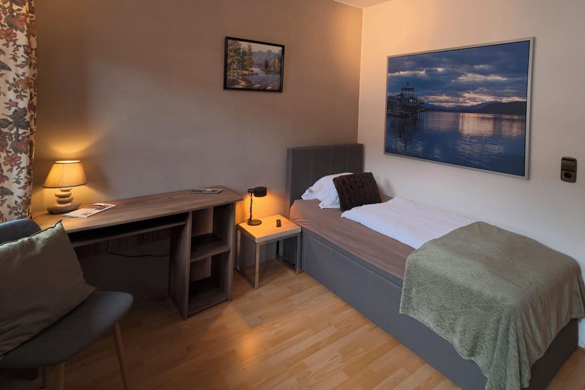 Une chambre d’hôtel de taille modeste, comprenant un lit simple, un bureau avec une chaise, une petite table avec une lampe et un tableau accroché au mur.