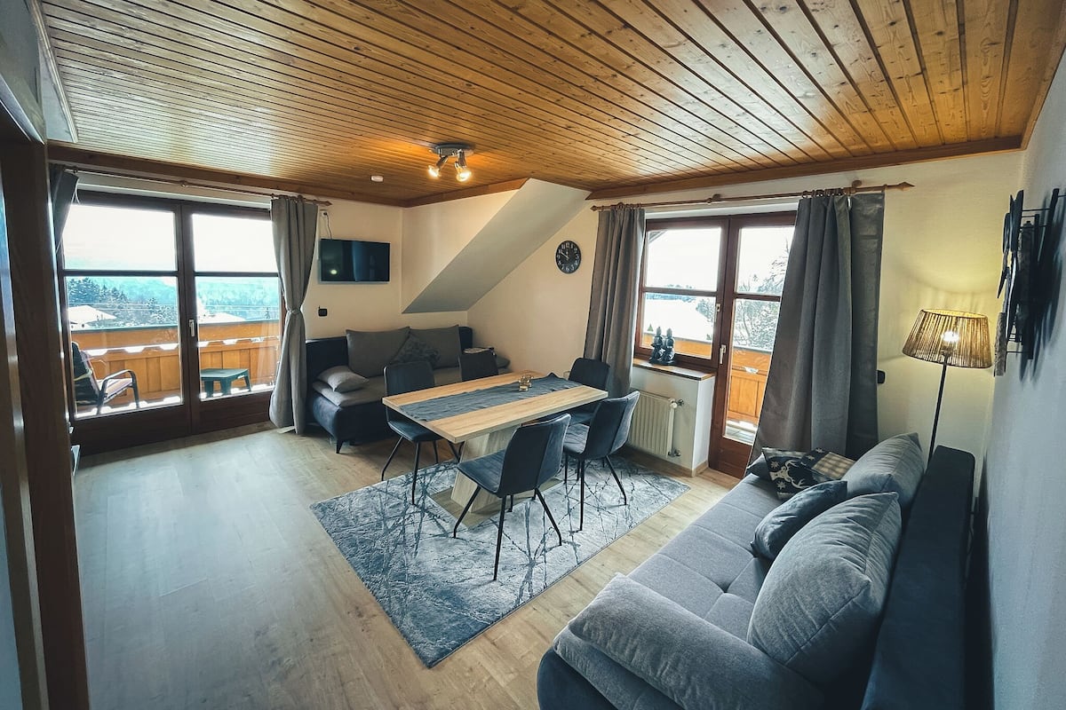 Un salon chaleureux avec un plafond en bois, un canapé, une table à manger avec des chaises et de grandes fenêtres donnant sur un beau paysage.