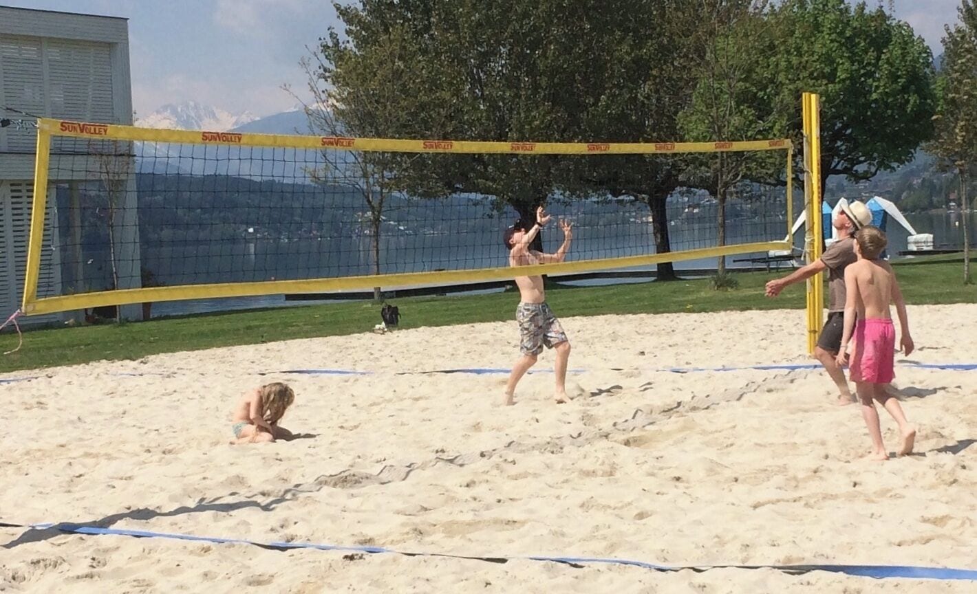 A sandy beach volleyball court with a net, trees, and a mountain in the background.