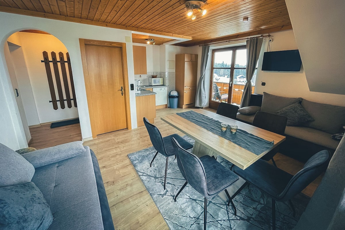 Un salon chaleureux avec un plafond en bois, un coin repas avec une table et des chaises, un canapé et un téléviseur fixé au mur.