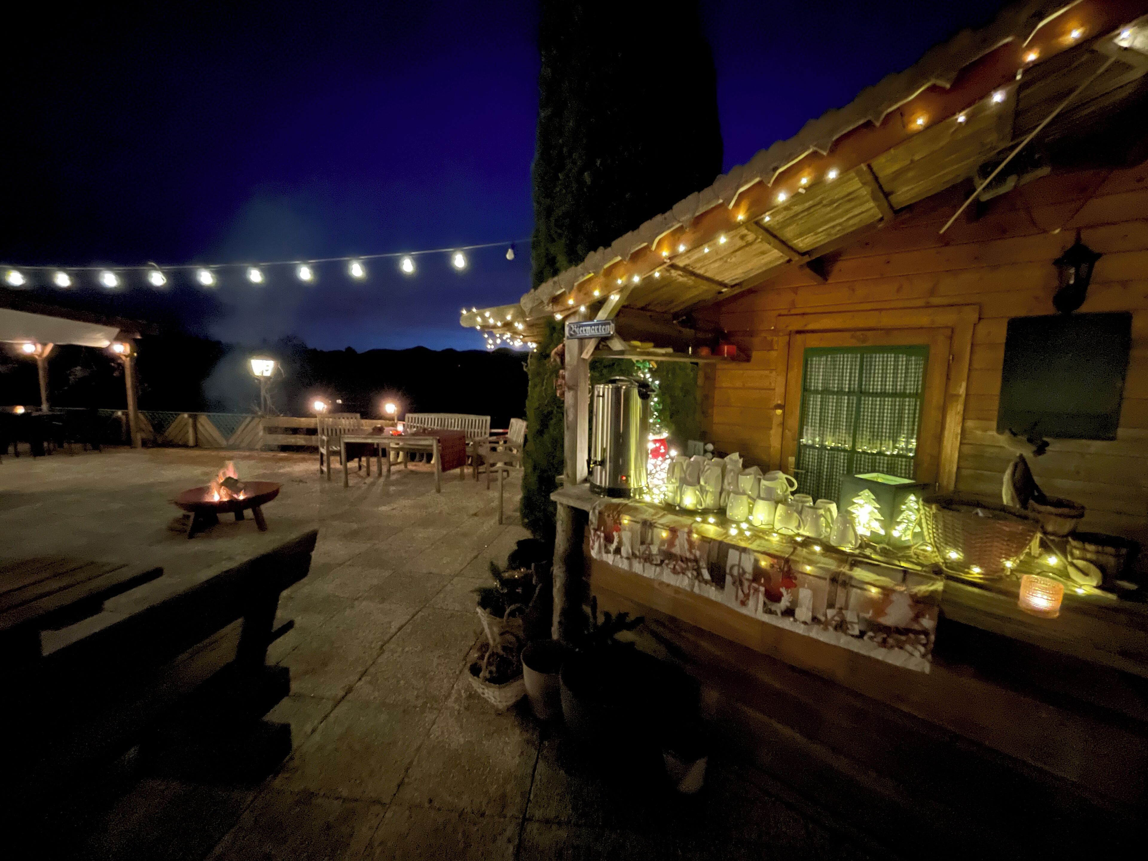 An outdoor seating area with string lights, a fire pit, and a bar area with bottles and glasses.