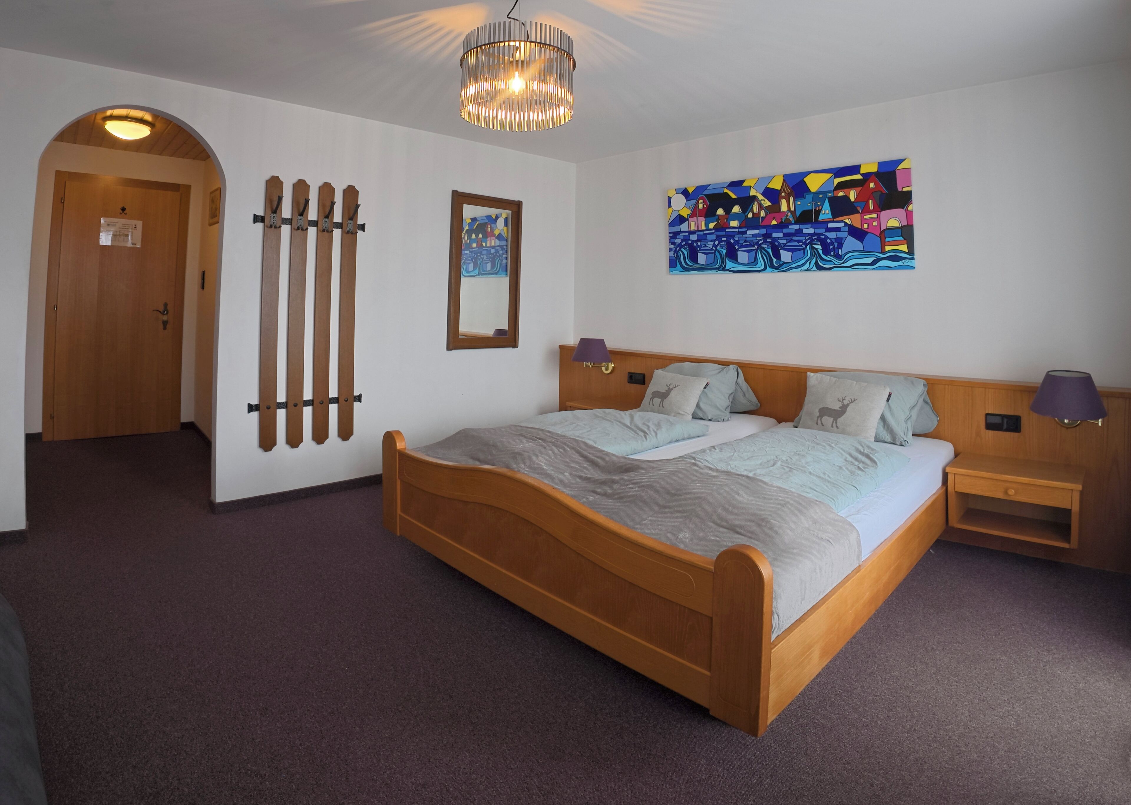 Une chambre à coucher avec un lit en bois, deux tables de chevet, une lampe fixée au mur, une porte, un porte-manteau et un tableau.