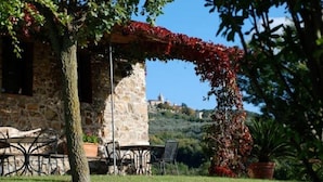 A stone building with a covered patio, outdoor seating, and a view of a hill with a building in the distance. - Agriturismo La Colombaia (Massa Marittima)