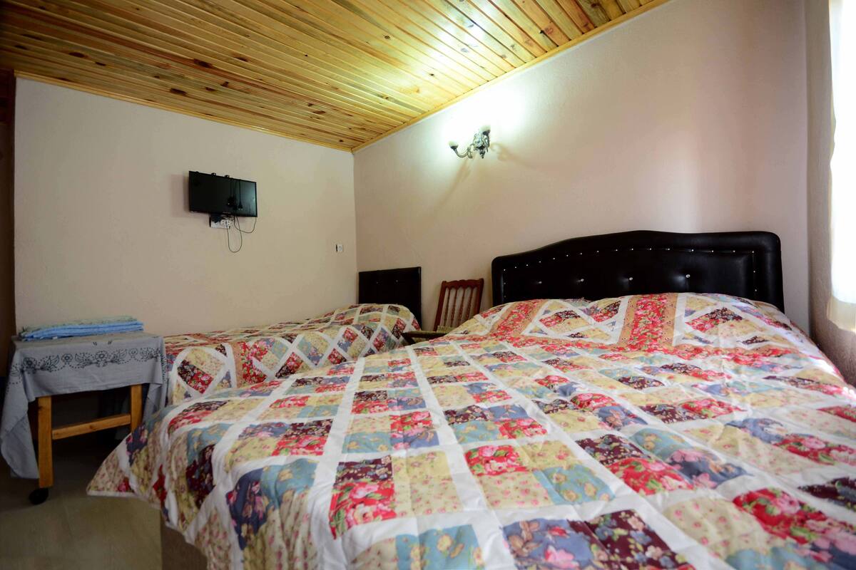 A bedroom with two beds, a wooden ceiling, a wall-mounted TV, and a small table with a tablecloth.