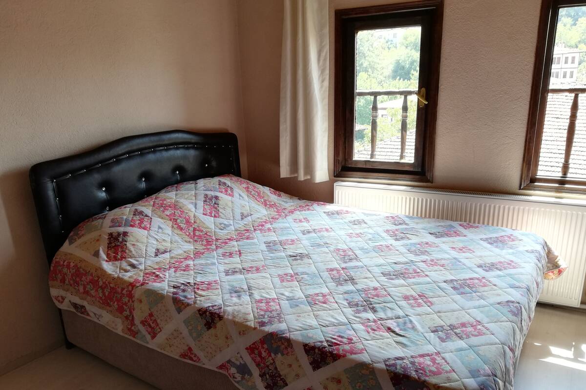 A bed with a floral quilt, a dark headboard, a window with a view of greenery, and a radiator.