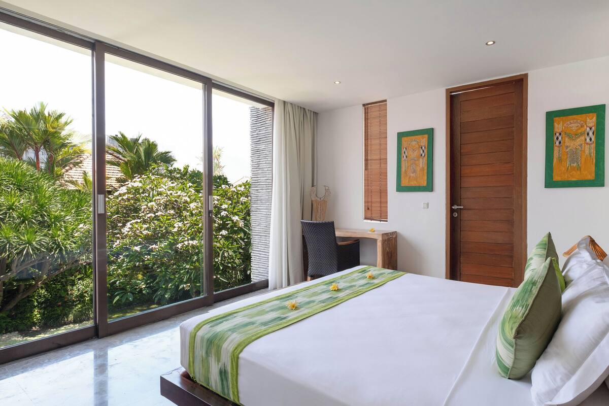 Un dormitorio moderno con una cama grande, un escritorio y vistas de exuberante vegetación a través de ventanas de pared a techo.
