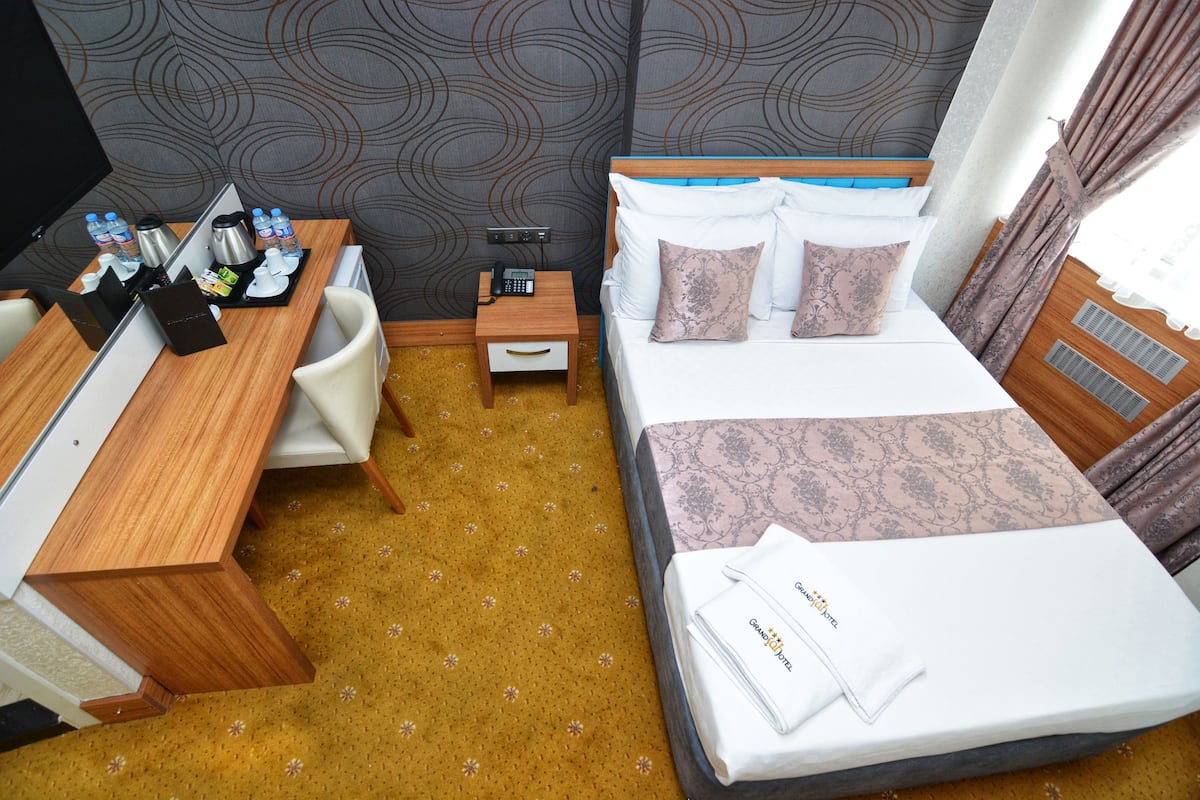 A hotel room with a bed, bedside table, and a desk with a chair. There is a small nightstand with a phone and a lamp. The room has a patterned wallpaper and a carpet with a floral design.