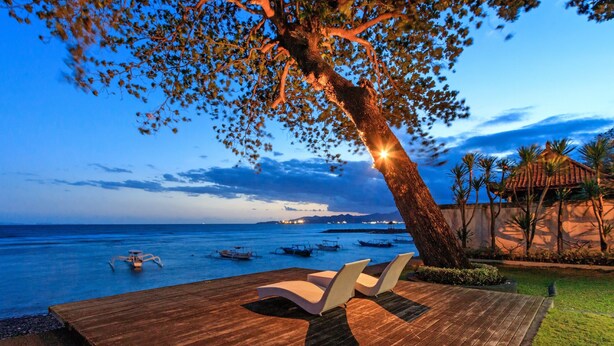 Una terraza de madera con dos sillas blancas, con vista a un mar tranquilo con barcos, un árbol y un edificio de techo rojo.