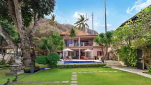 Una casa de dos pisos con piscina, rodeada de exuberante follaje y palmeras.