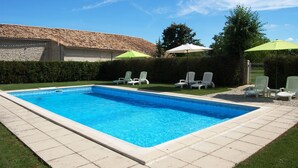 Una piscina rectangular con un borde empedrado, rodeada de sillas de descanso blancas y sombrillas verdes.
