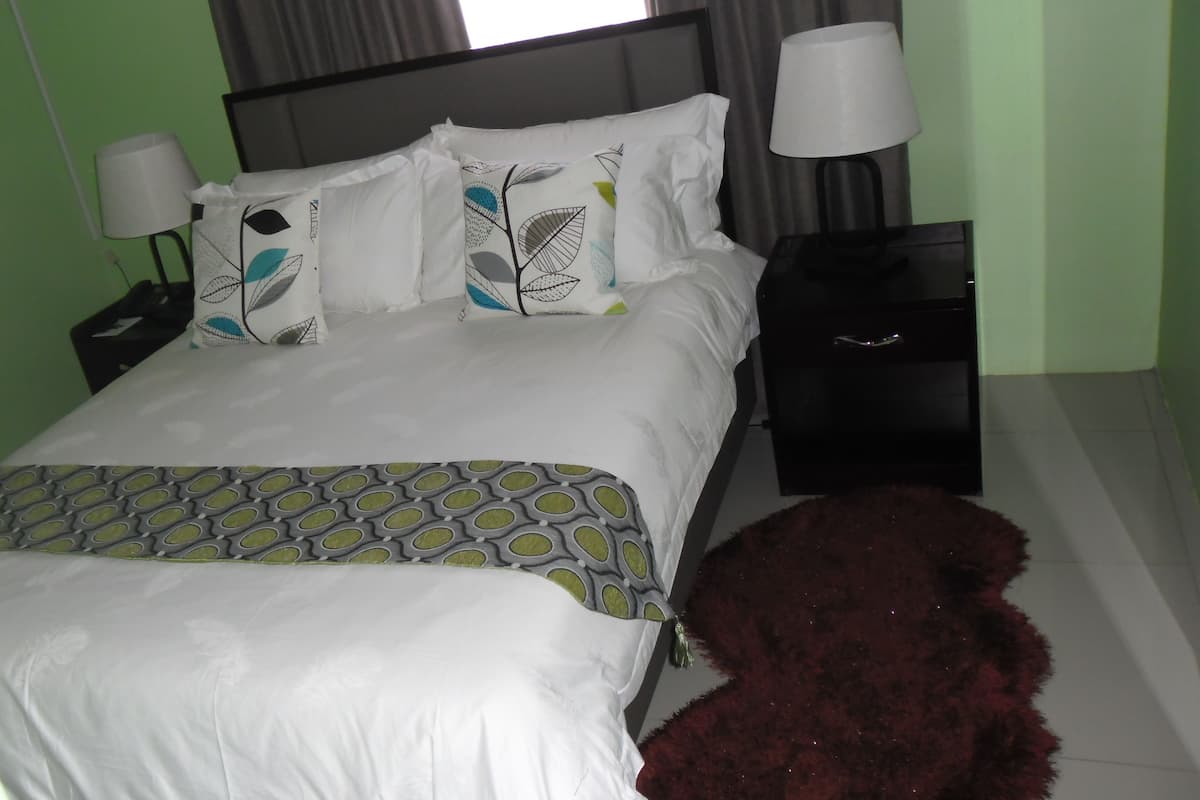 A neatly made bed with patterned pillows and a matching throw, a nightstand with a lamp, and a red furry rug beside the bed.
