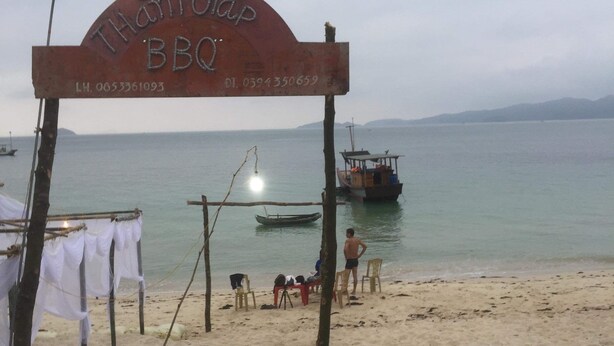 Un panneau indiquant qu’il y a un barbecue sur la plage, avec des numéros de téléphone à contacter, une plage de sable, quelques personnes et un bateau en arrière-plan.