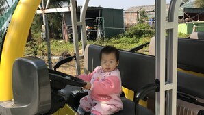 Un enfant est assis dans un véhicule jaune doté d’un toit, qui pourrait être une petite voiture ou un véhicule de golf, dans un endroit en plein air, avec des arbres et un bâtiment en arrière-plan.