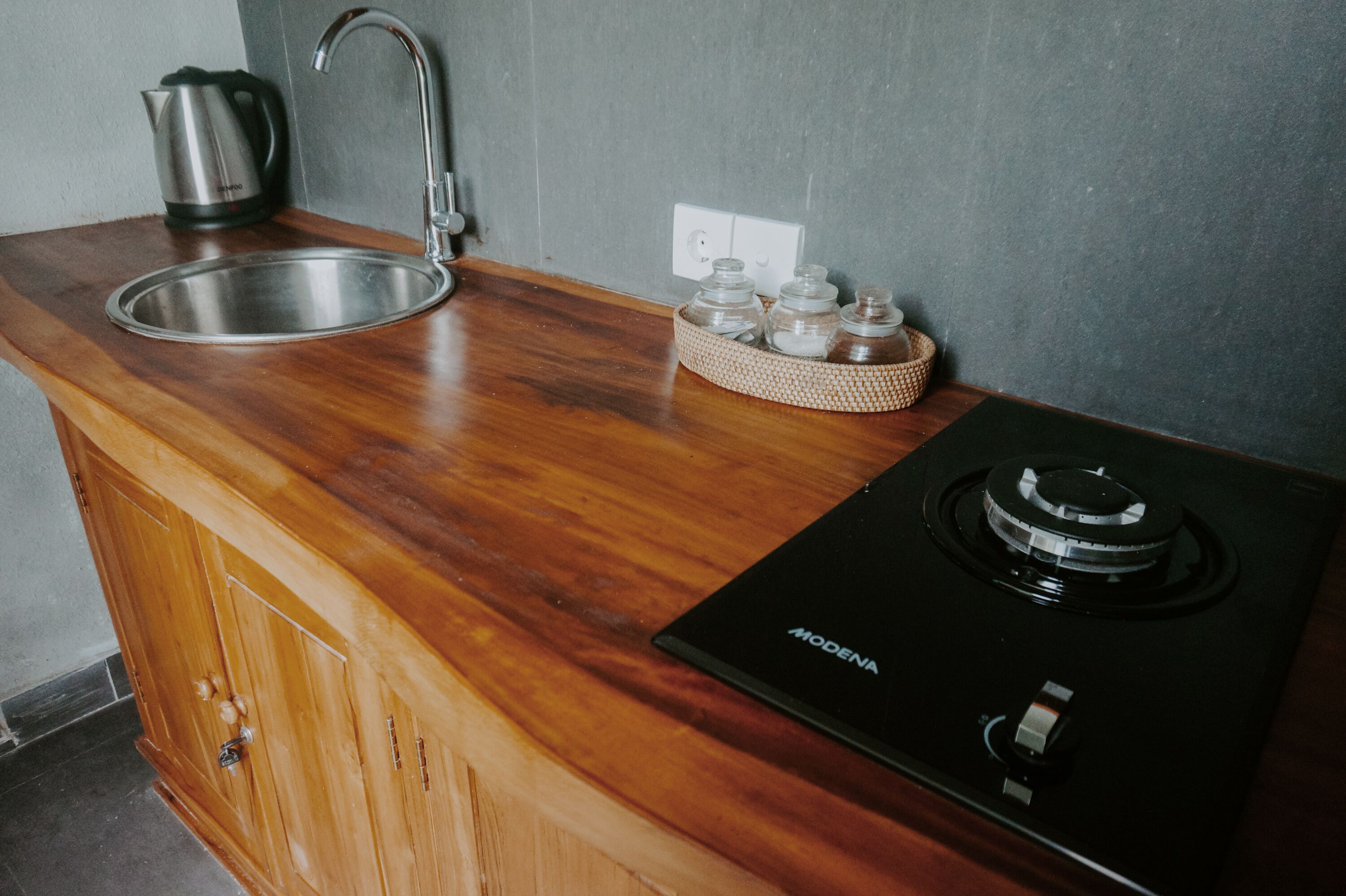 A kitchen with a stainless steel sink, a kettle, a stove, and a woven basket with jars.