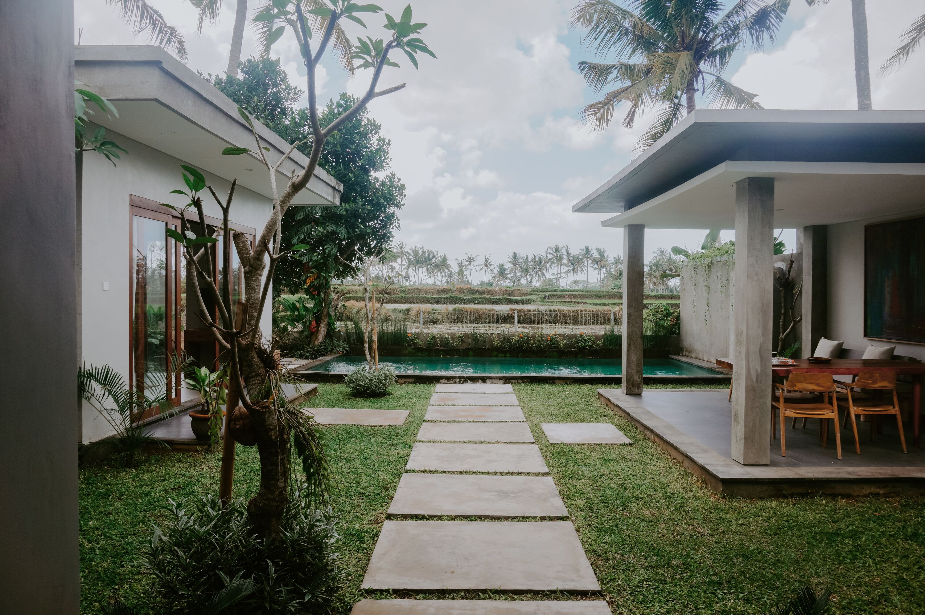A modern house with a concrete pathway leading to a pool, surrounded by lush greenery and palm trees.
