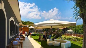 An outdoor seating area with tables and chairs, a covered seating area with umbrellas, and a landscaped garden with a tree and flowers. - Guest House Terra di Venera (Acireale)
