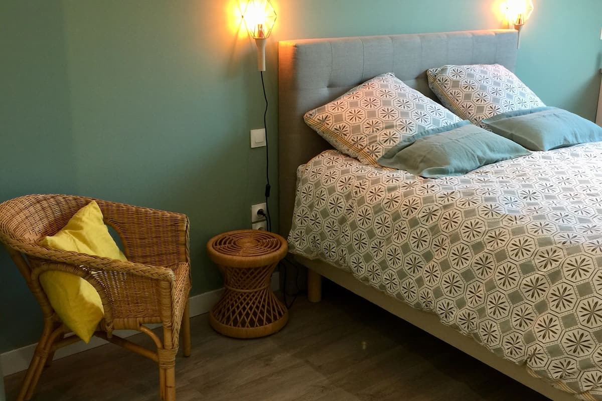 Un dormitorio con una cama, dos lámparas fijadas a la pared, una silla de mimbre con cojín y un taburete de madera.