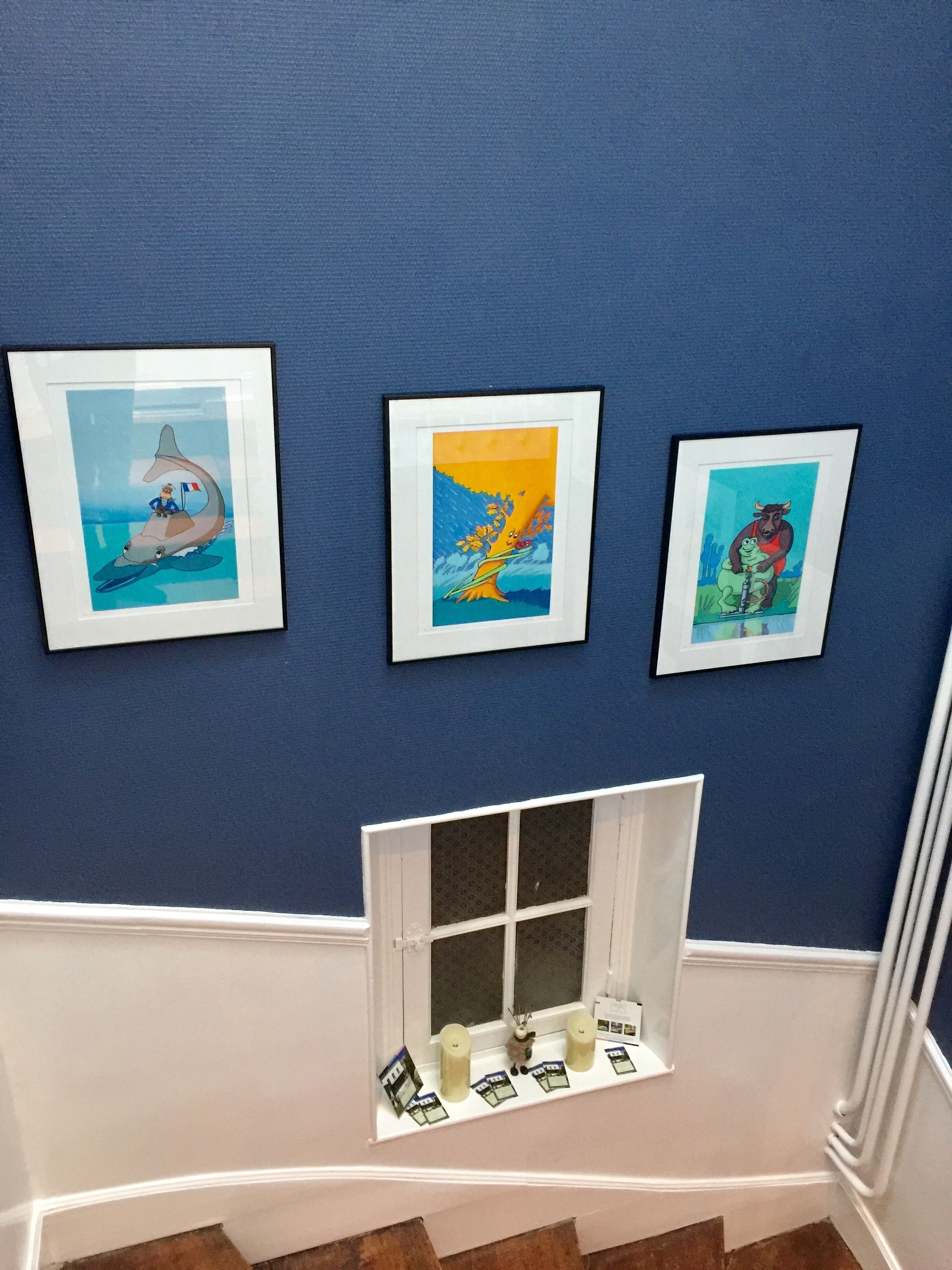 Three framed artworks on a blue wall, a window with candles and cards, and a white window frame.