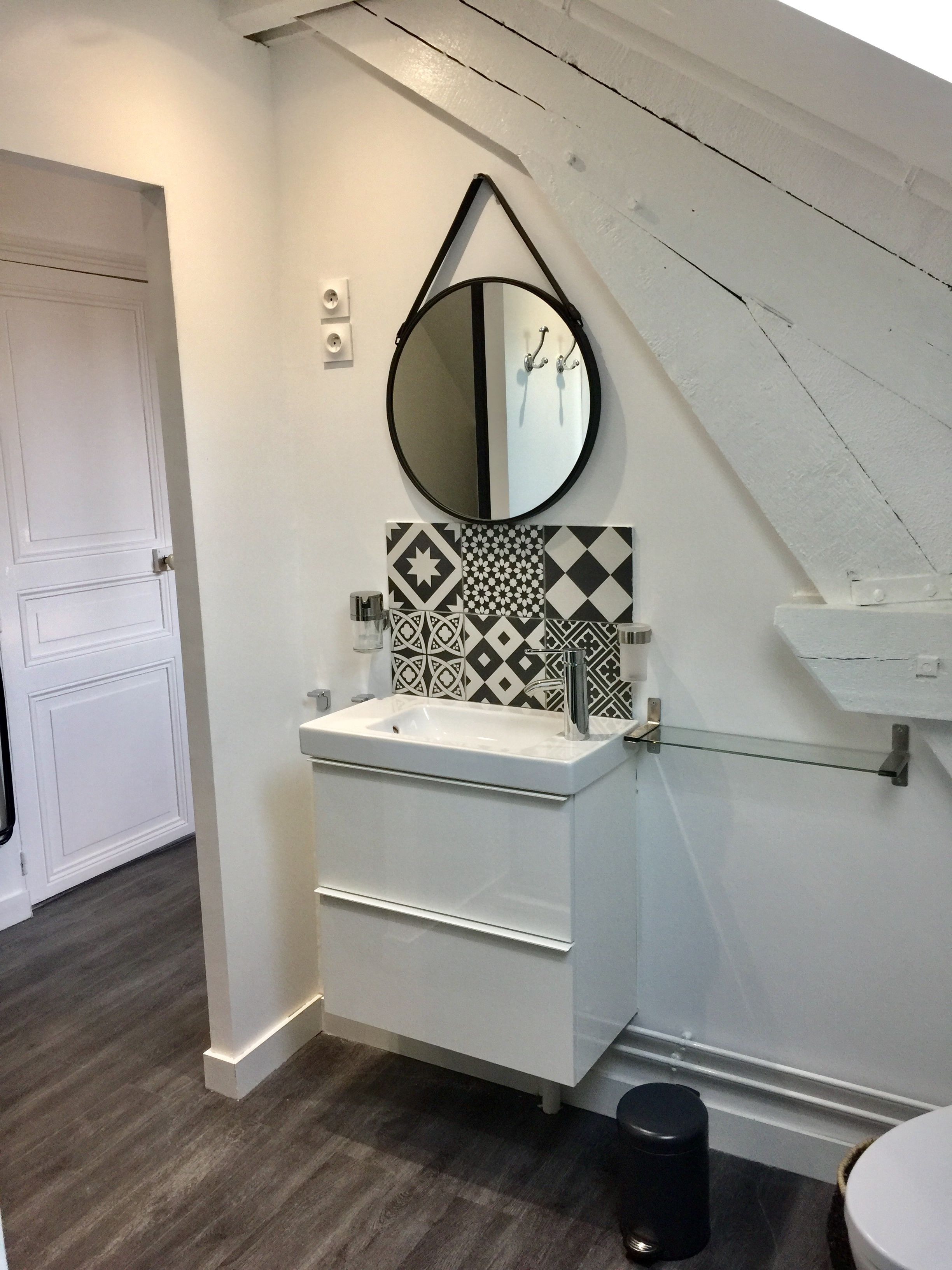 A bathroom with a white vanity, a round mirror, patterned tiles, and a black trash bin.