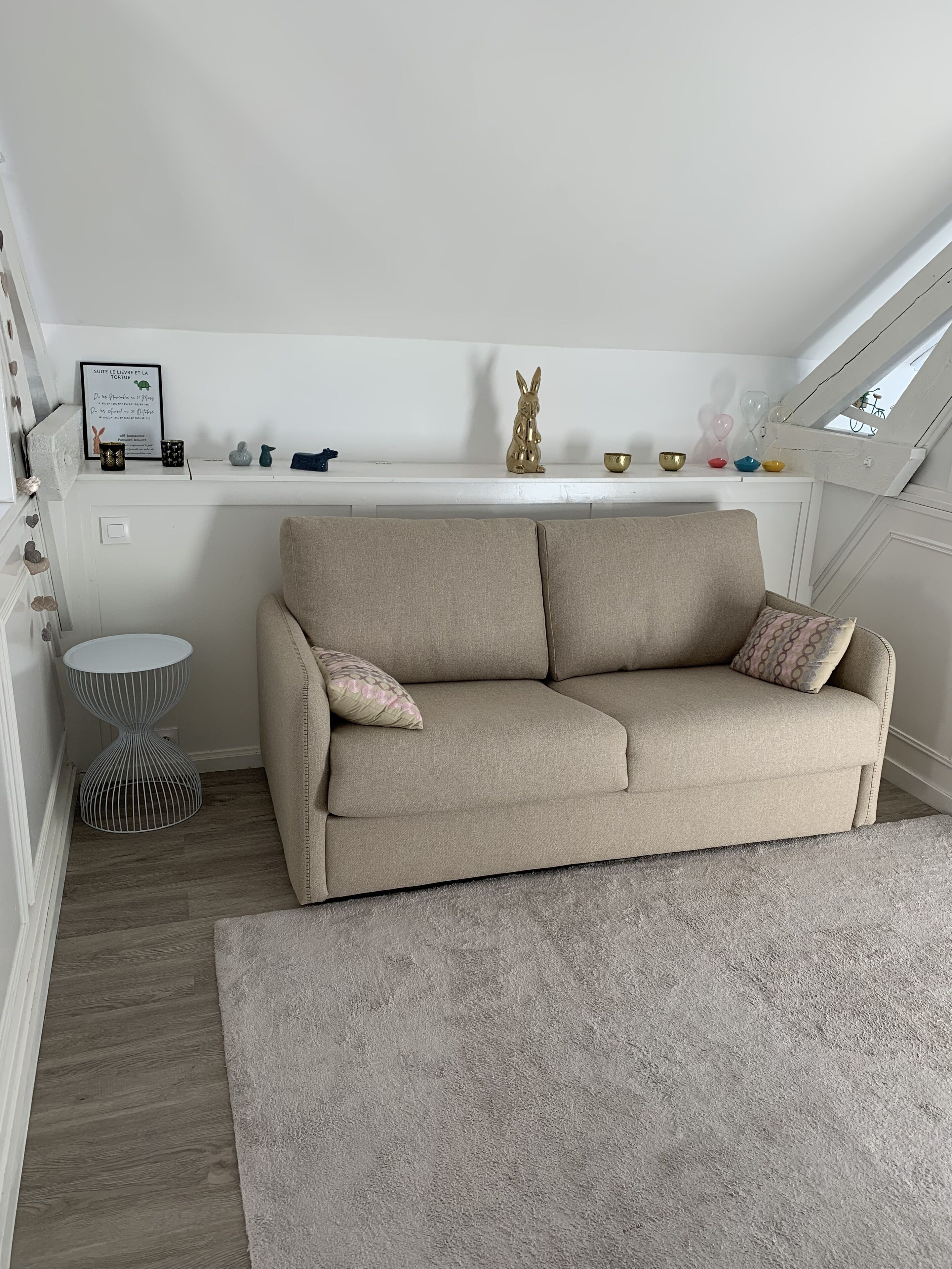 Een knus loft slaapkamer met een beige bank, een klein rond tafeltje en een decoratieve plank met diverse spullen.