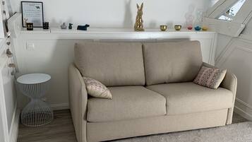 Een knus loft slaapkamer met een beige bank, een klein rond tafeltje en een decoratieve plank met diverse spullen.