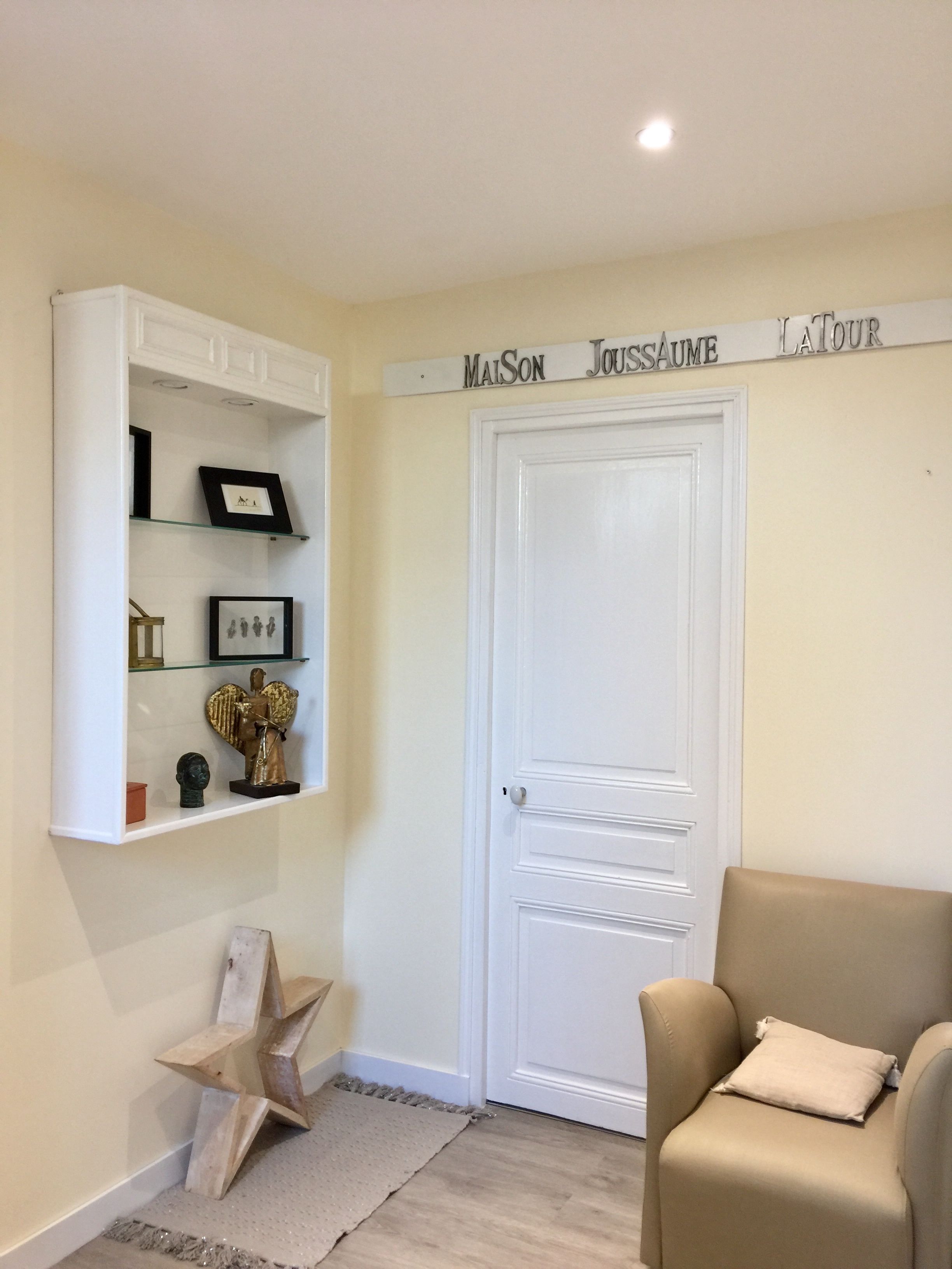 Una habitación con una puerta blanca, un sillón color crema, un adorno con forma de estrella y una estantería con marcos y una estatua dorada de un ángel.