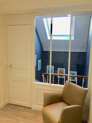 Una habitación con una puerta blanca, una ventana con barandilla de madera y un sillón color crema.
