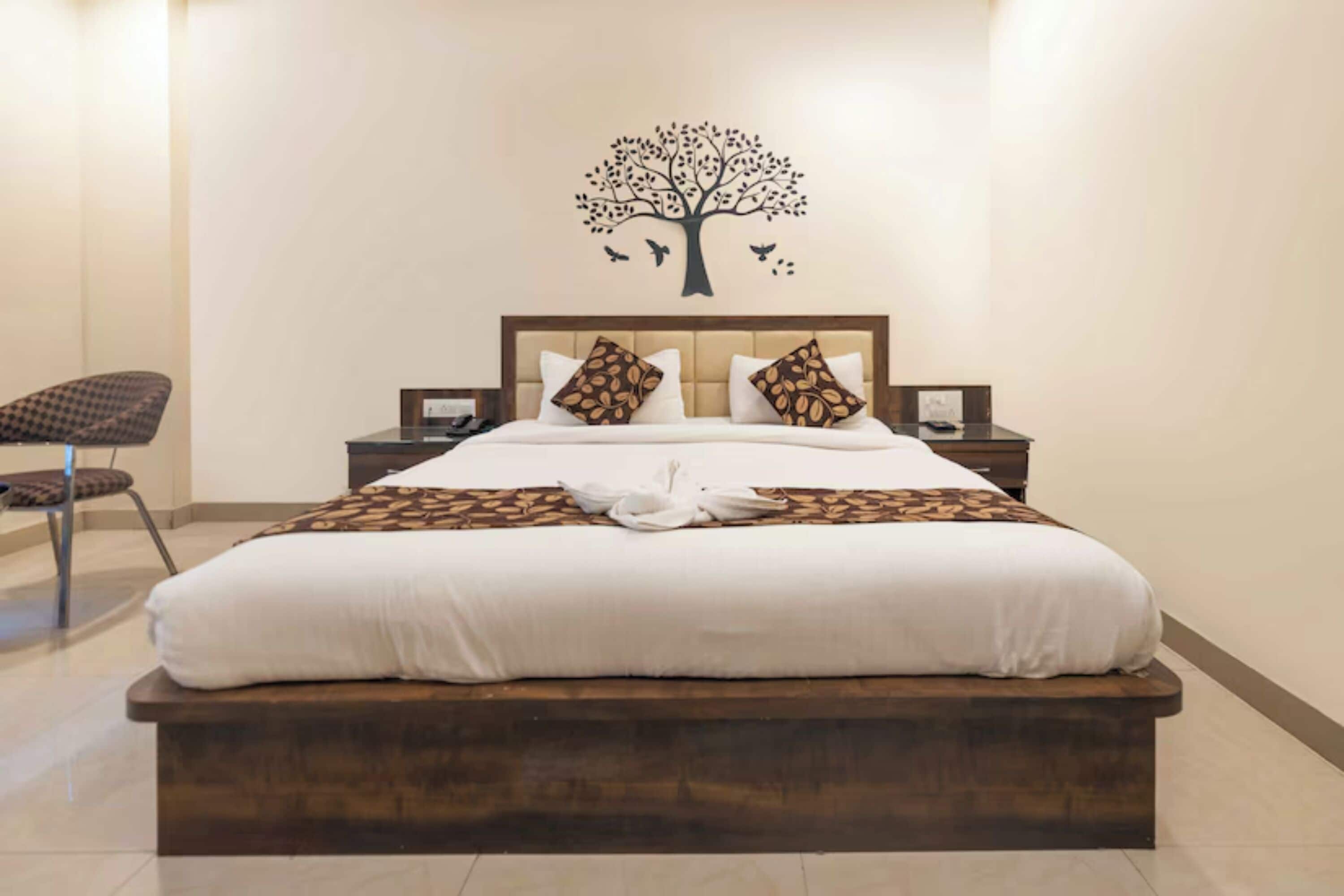 A hotel room with a large bed, a wooden headboard, two nightstands, a chair, and a wall decoration of a tree with birds.