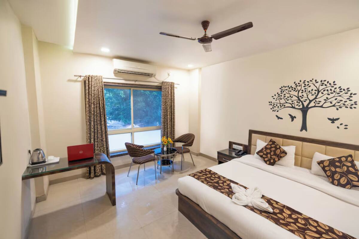Quarto de hotel com uma cama, escrivaninha, cadeira, mesa, ventoinha no teto e janela com cortinas.