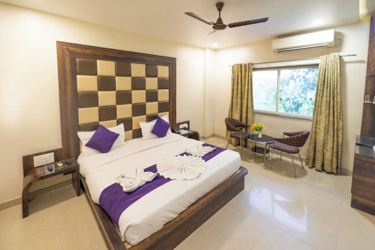 Quarto de hotel com cabeceira xadrez, cama com roupa de cama branca e roxa, uma mesa pequena com cadeiras e uma janela com cortinas.