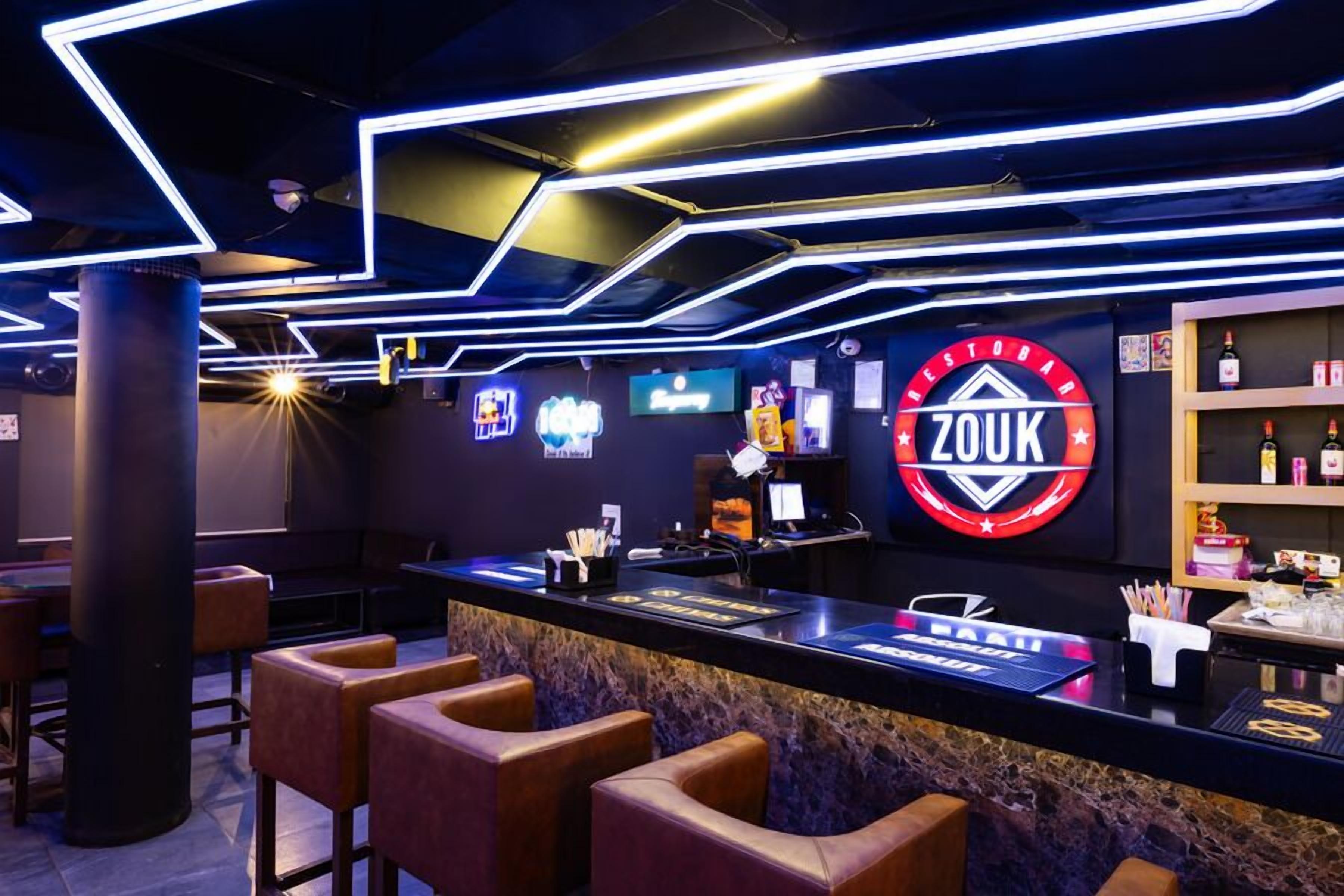 Zona de bar con un letrero de neón que dice “ZOUK”, taburetes, una barra con menú y una estantería con botellas.