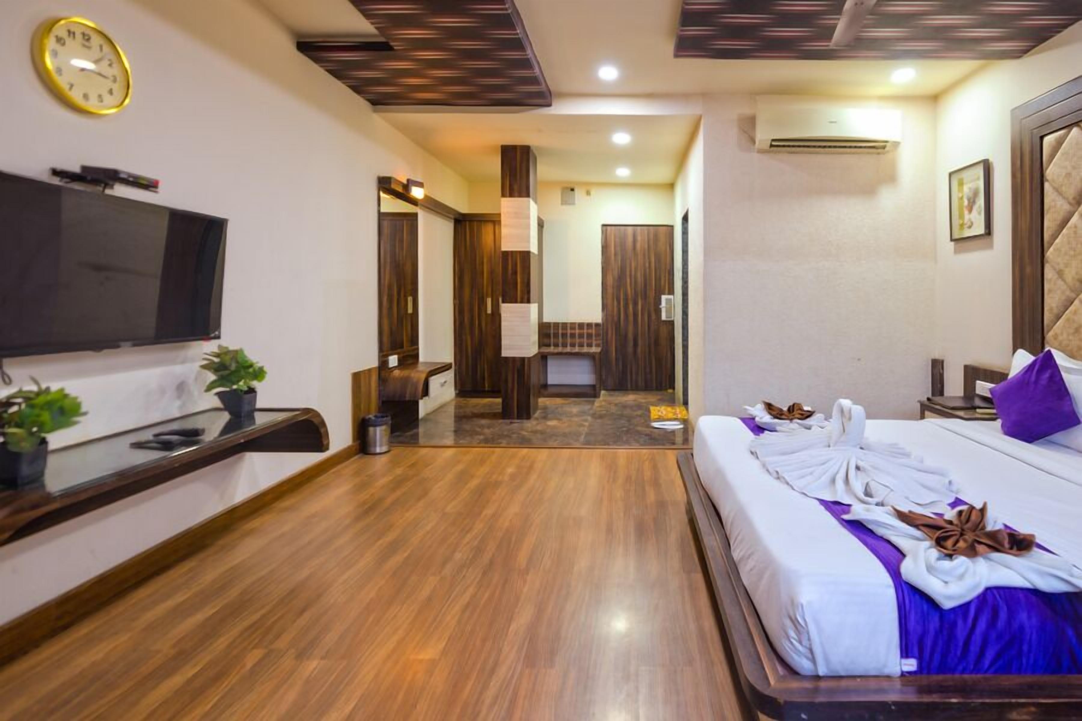 Una camera d'hotel con pavimento in legno, televisione a schermo piatto, un letto con biancheria da letto bianca e viola, e un bagno visibile attraverso una porta aperta.