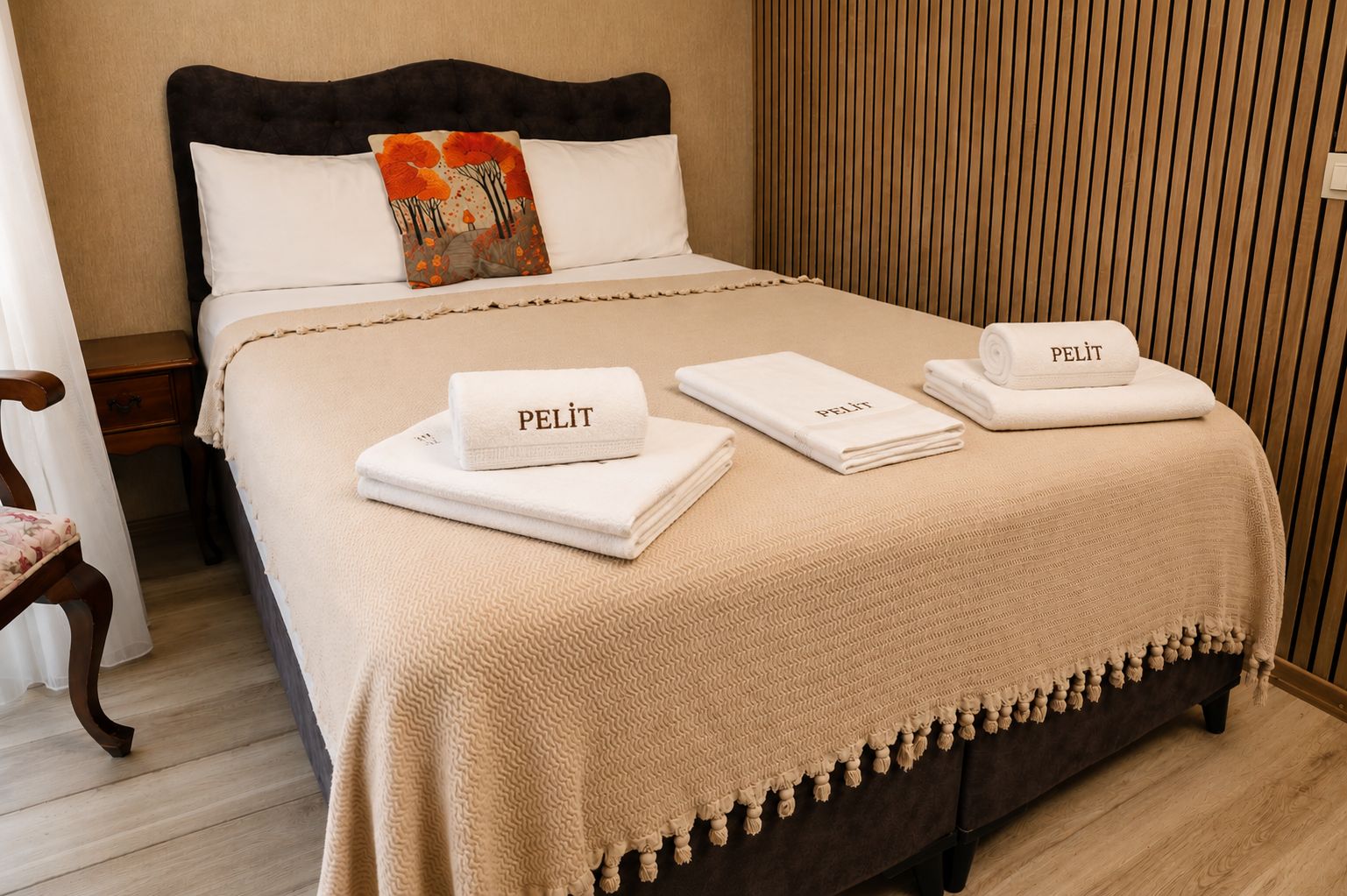 Una cama bien tendida con cabecera oscura, almohadas blancas y una almohada decorativa con una escena de naturaleza. Hay tres toallas dobladas con la palabra 'PELIT' y una manta beige texturizada con flecos al pie de la cama.